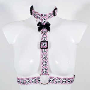May include: Pink and black harness with skull and crossbones print and a black bow. The harness has adjustable straps and a metal ring.