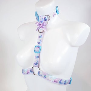 May include: A white harness with a light blue and purple moon and star pattern. The harness has a matching light blue choker with a purple bow and a silver ring.