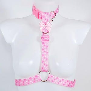 May include: Pink pet harness with a paw print pattern. The harness includes a collar, chest strap, and waist straps, all connected with metal rings and adjustable buckles. A small pink bow is attached to the chest strap. The harness is displayed on a white mannequin.