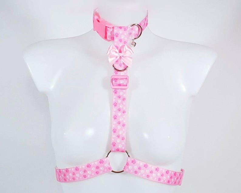 May include: Pink pet harness with a paw print pattern. The harness includes a collar, chest strap, and waist straps, all connected with metal rings and adjustable buckles. A small pink bow is attached to the chest strap. The harness is displayed on a white mannequin.