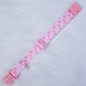 May include: Pink pet collar with white paw prints, a plastic buckle, and a metal bell. The collar is made of a durable fabric and features a quick-release buckle for easy use. A charming accessory for your furry friend.