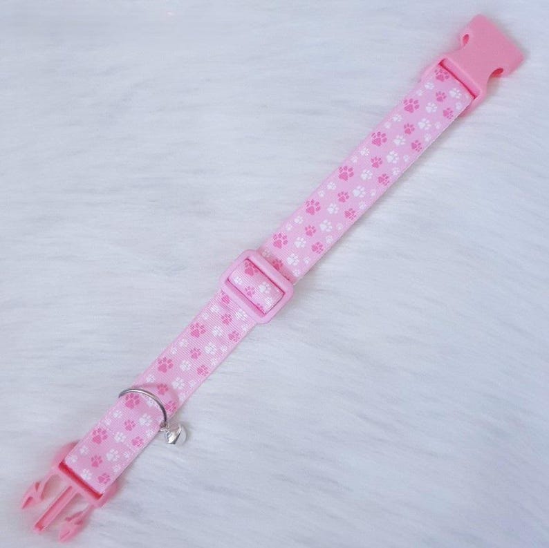 May include: Pink pet collar with white paw prints, a plastic buckle, and a metal bell. The collar is made of a durable fabric and features a quick-release buckle for easy use. A charming accessory for your furry friend.