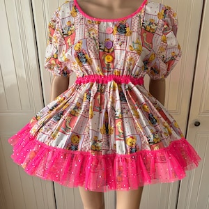 Op de afbeelding: Een witte jurk met een roze en witte teddybeerprint. De jurk heeft een roze elastische tailleband en een roze tule ruche met roze glitter.