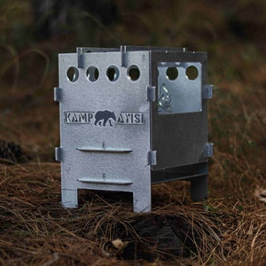 Puede incluir: Una estufa de camping de metal plateado con un logotipo de oso que dice "Kamp Ayisi". La estufa tiene una pequeña ventana en el lateral y algunos agujeros en la parte delantera.
