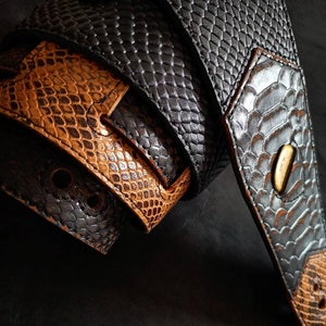 Puede incluir: Primer plano de dos correas de guitarra con un patrón de piel de serpiente. Una correa es negra y la otra marrón. Las correas están hechas de cuero y tienen una superficie texturizada. Las correas están entrelazadas, mostrando los detalles de las costuras y los herrajes.