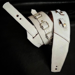 May include: Two white leather guitar straps with silver buckles and stitching. One strap has a single buckle and the other has three buckles.
