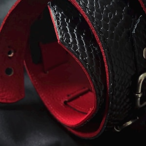 May include: A black belt with a textured snakeskin pattern on the exterior and a vibrant red interior lining. The belt features a metallic buckle and visible adjustment holes along the strap, showcasing a high-contrast colour combination.