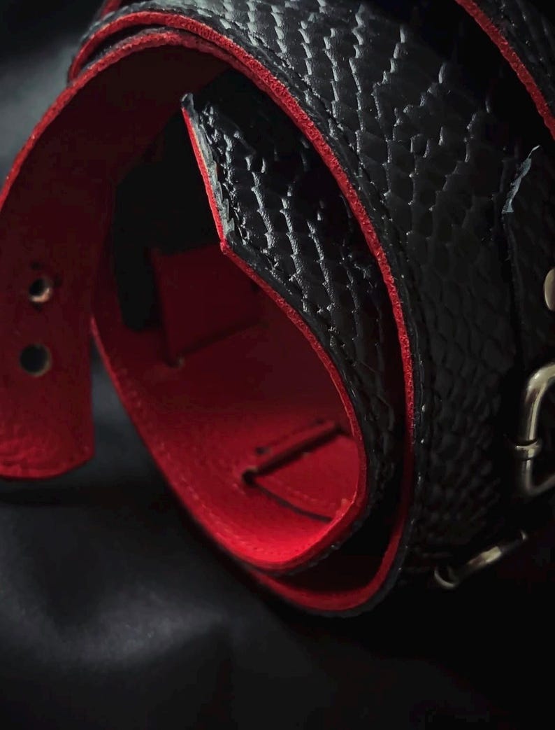 May include: A black belt with a textured snakeskin pattern on the exterior and a vibrant red interior lining. The belt features a metallic buckle and visible adjustment holes along the strap, showcasing a high-contrast colour combination.