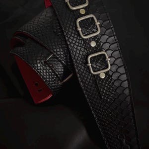 May include: A black belt featuring a textured snakeskin pattern and a bright red underside. The belt is adorned with three silver-toned square buckles arranged vertically along the strap, secured by silver-toned metal rivets.