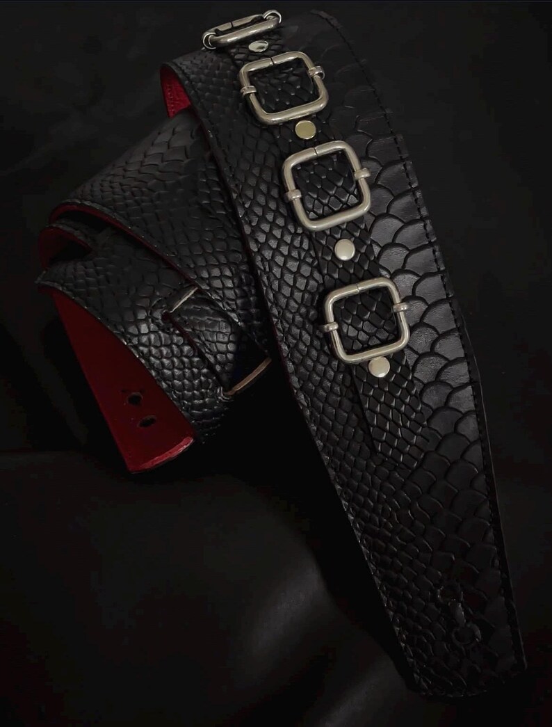 May include: A black belt featuring a textured snakeskin pattern and a bright red underside. The belt is adorned with three silver-toned square buckles arranged vertically along the strap, secured by silver-toned metal rivets.
