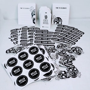 May include: A collection of black and white stickers featuring a sugar skull design. The stickers include various sizes and shapes, including round stickers with the text "Hello I've Arrived" and rectangular stickers with the text "Leave Your Mark". There are also gift tags with the text "Ceci Ruiz" and note cards with the text "Time to Celebrate".