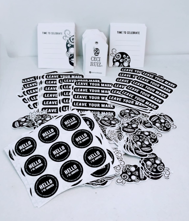 May include: A collection of black and white stickers featuring a sugar skull design. The stickers include various sizes and shapes, including round stickers with the text "Hello I've Arrived" and rectangular stickers with the text "Leave Your Mark". There are also gift tags with the text "Ceci Ruiz" and note cards with the text "Time to Celebrate".