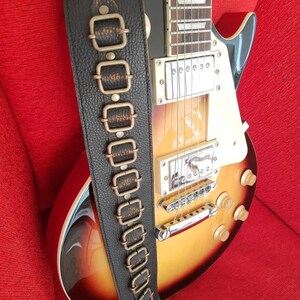 May include: A brown leather guitar strap with decorative metal accents. The strap has a textured surface and is adorned with multiple rows of metal buckles.