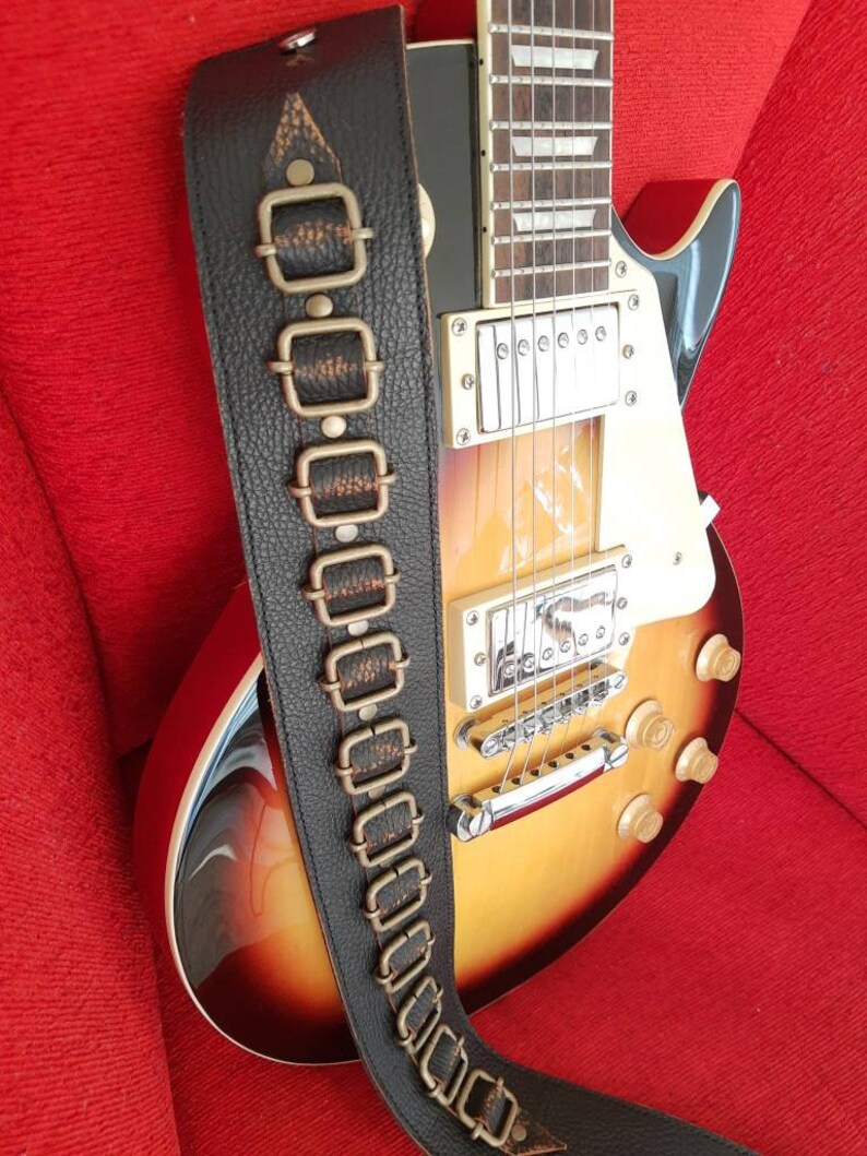May include: A brown leather guitar strap with decorative metal accents. The strap has a textured surface and is adorned with multiple rows of metal buckles.