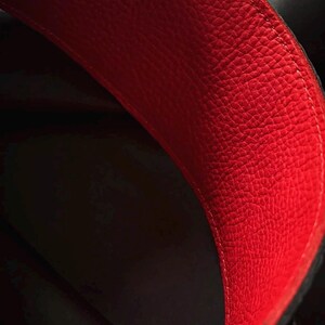 May include: A close-up view of the interior of a bag featuring a vibrant red, textured leather lining. The material displays a distinct pebbled grain pattern with visible stitching along the curved edge, contrasting against the dark, shadowed depth.