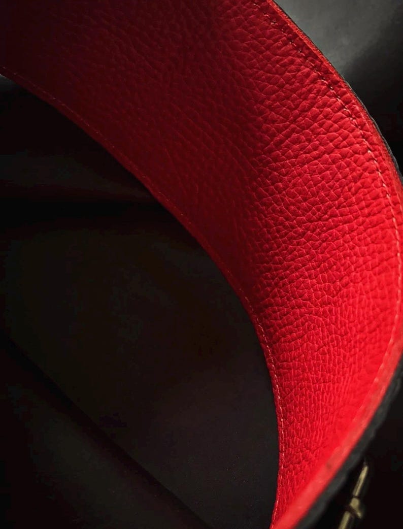 May include: A close-up view of the interior of a bag featuring a vibrant red, textured leather lining. The material displays a distinct pebbled grain pattern with visible stitching along the curved edge, contrasting against the dark, shadowed depth.