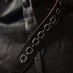 May include: A black guitar strap featuring a textured snakeskin pattern on the exterior and a vibrant red lining on the interior. The strap is adorned with a series of silver-toned metal rectangular buckles and rivets along its length.