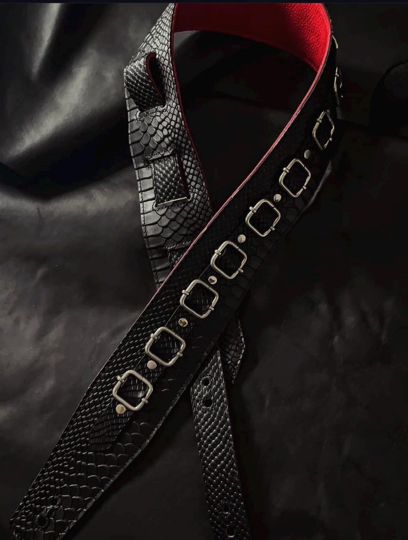 May include: A black guitar strap featuring a textured snakeskin pattern on the exterior and a vibrant red lining on the interior. The strap is adorned with a series of silver-toned metal rectangular buckles and rivets along its length.