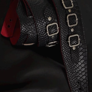 May include: A black guitar strap featuring a textured snakeskin pattern on the exterior and a solid red lining on the interior. The strap includes three silver-toned metal buckles and rivets, with reinforced holes at the end for attachment.