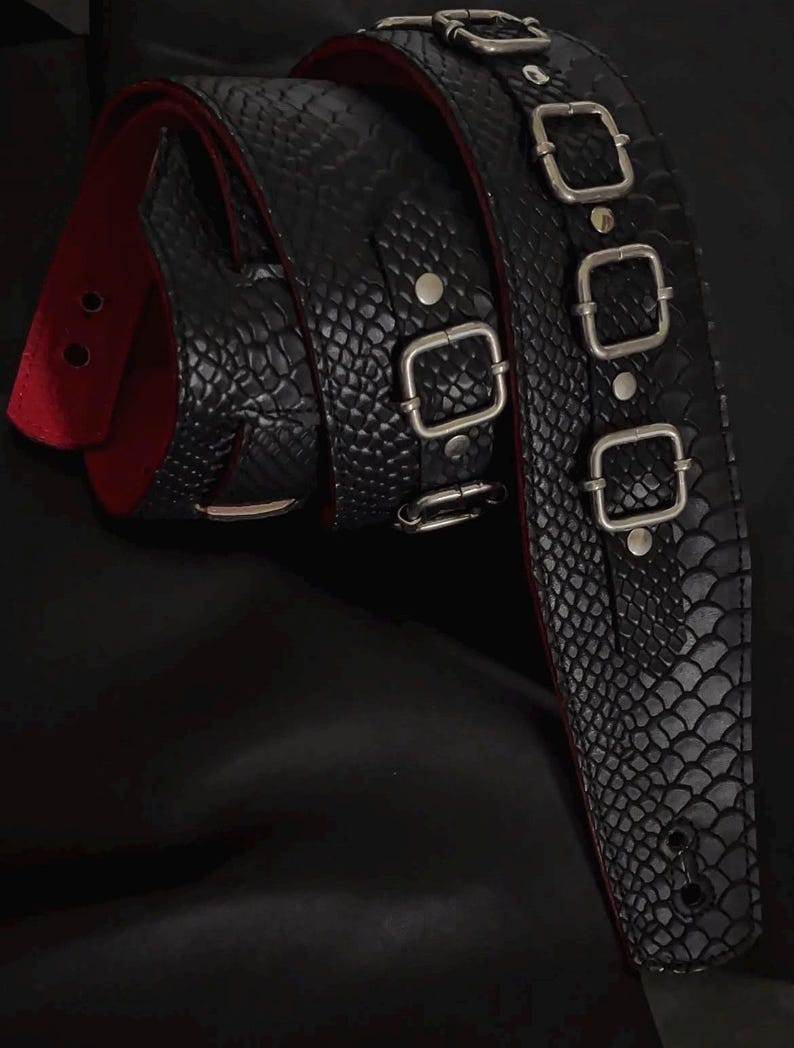 May include: A black guitar strap featuring a textured snakeskin pattern on the exterior and a solid red lining on the interior. The strap includes three silver-toned metal buckles and rivets, with reinforced holes at the end for attachment.