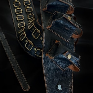 May include: Black leather holster with multiple pouches and brass-colored buckles. The holster has a textured surface and is stitched with white thread. A smaller strap and a decorative patch with a skull are also visible.