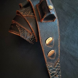 Pode incluir: Uma correia de guitarra de couro preto com costuras marrons e três detalhes em metal dourado. A correia tem um pequeno bolso na parte superior para um palheta ou outro objeto pequeno.