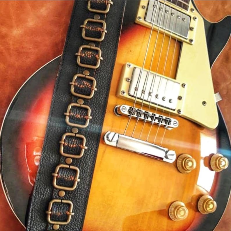 May include: A close-up of a vintage-style electric guitar with a sunburst finish. The guitar has a black leather strap with brass accents. The guitar has a gold-colored pickguard and chrome hardware.