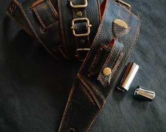 May include: A black textured leather guitar strap featuring antique brass buckles, adjustment loops, and a small attached pouch with a brass snap closure. Two polished silver metal guitar slides are positioned next to the strap.