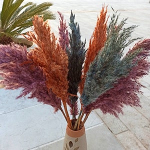 May include: A bouquet of dried pampas grass in a variety of colors, including pink, orange, gray, and blue, arranged in a ceramic vase with a leaf design.
