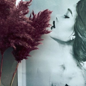 May include: A black and white photograph of a woman's face with her eyes closed and lips painted black. A sprig of dried, burgundy pampas grass is in the foreground.