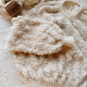 May include: A cream-colored knitted sweater with a textured pattern. The sweater is laid out flat on a light gray surface. A ball of cream-colored yarn is in the background with a label that reads "The Wave Wool".