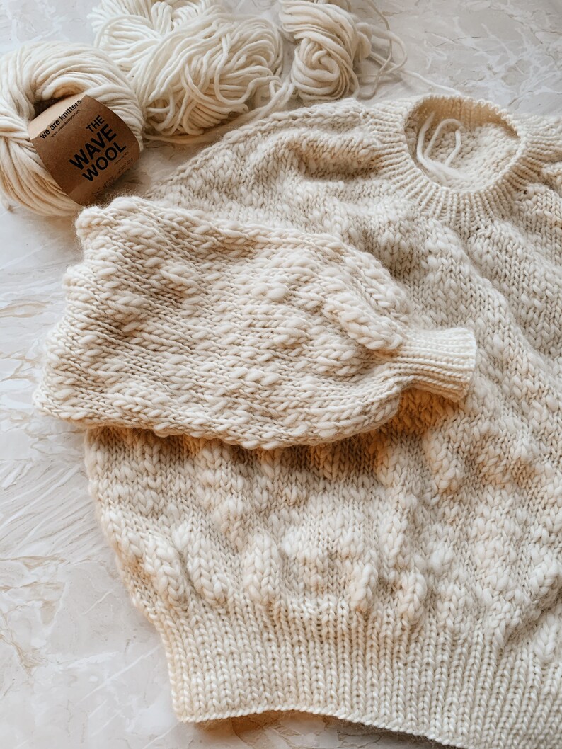 May include: A cream-colored knitted sweater with a textured pattern. The sweater is laid out flat on a light gray surface. A ball of cream-colored yarn is in the background with a label that reads "The Wave Wool".