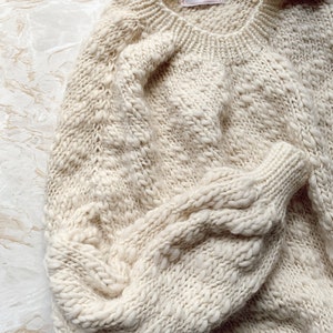 May include: A cream-colored knitted sweater with a cable knit pattern. The sweater has a round neckline and long sleeves. The tag on the sweater reads "KnitsByCari."