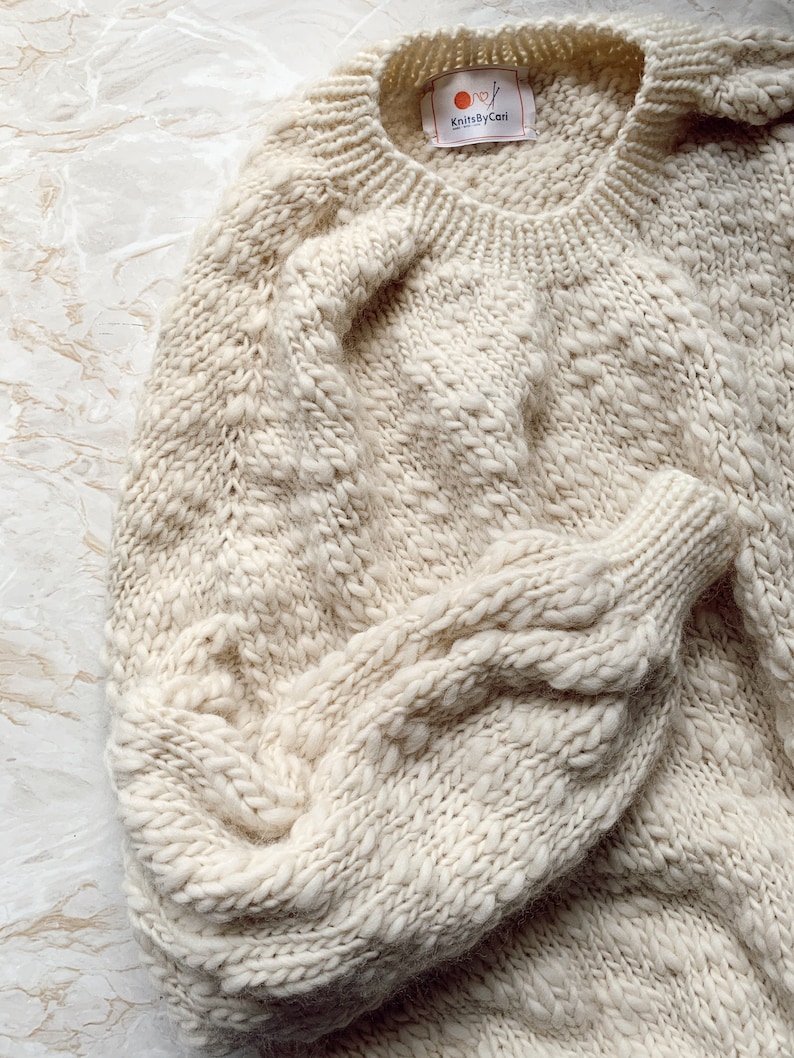 May include: A cream-colored knitted sweater with a cable knit pattern. The sweater has a round neckline and long sleeves. The tag on the sweater reads "KnitsByCari."