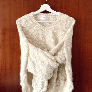 May include: A cream-colored knitted sweater with a cable knit pattern hangs on a white hanger against a wood background. The sweater has a round neckline and long sleeves.