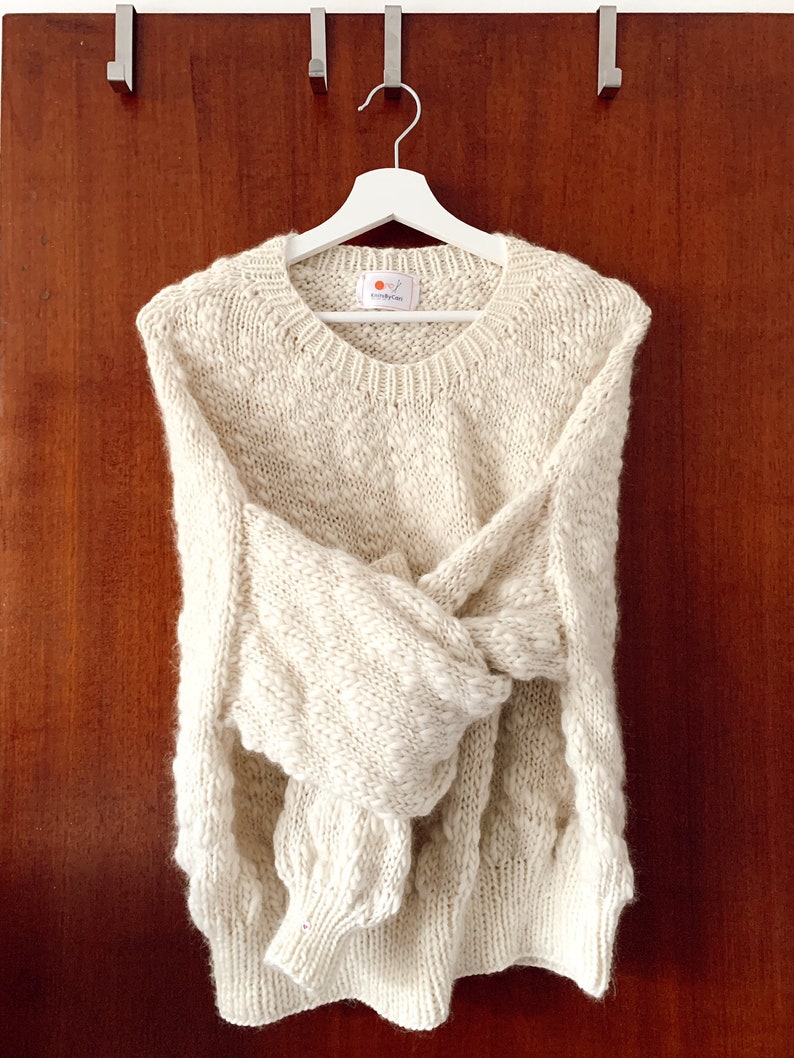 May include: A cream-colored knitted sweater with a cable knit pattern hangs on a white hanger against a wood background. The sweater has a round neckline and long sleeves.