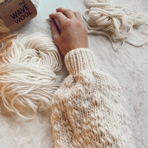 May include: A close-up of a white knitted sweater with a textured pattern. The sweater is made with a thick yarn and has a loose, oversized fit. There are two balls of white yarn in the background, one with a label that reads "The Wave Wool".