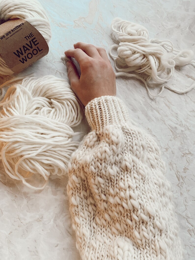 May include: A close-up of a white knitted sweater with a textured pattern. The sweater is made with a thick yarn and has a loose, oversized fit. There are two balls of white yarn in the background, one with a label that reads "The Wave Wool".