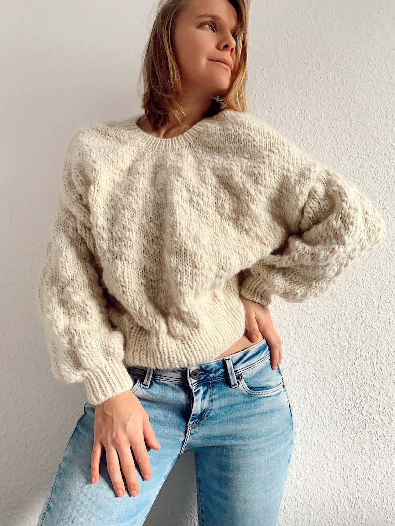 May include: A cream-colored knitted sweater with a textured pattern. The sweater has a round neckline and long sleeves. The model is wearing blue jeans.