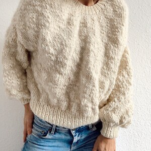 May include: A cream-colored knitted sweater with a textured pattern. The sweater has a round neckline and long sleeves.