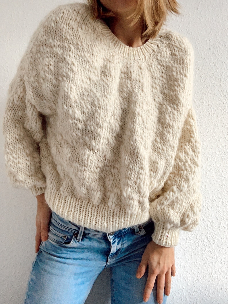 May include: A cream-colored knitted sweater with a textured pattern. The sweater has a round neckline and long sleeves.