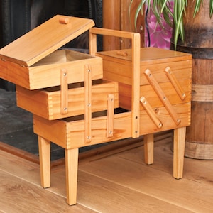 May include: A light brown wooden sewing box with a cantilevered design featuring multiple tiered compartments that extend outwards. The box stands on four tapered legs and includes a central handle, with visible finger joints on the corners.