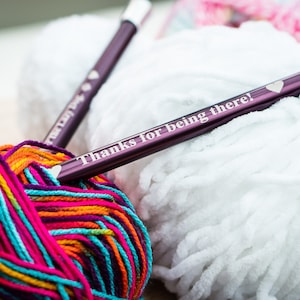 Pu&ograve; includere: Due ferri da maglia viola con un design a forma di cuore e il testo "Grazie per essere l&igrave;!" su uno dei ferri. I ferri sono appoggiati su un gomitolo di lana bianca e un gomitolo di lana multicolore.