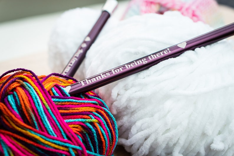 Pu&ograve; includere: Due ferri da maglia viola con un design a forma di cuore e il testo "Grazie per essere l&igrave;!" su uno dei ferri. I ferri sono appoggiati su un gomitolo di lana bianca e un gomitolo di lana multicolore.