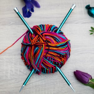 Pu&ograve; includere: Un gomitolo di lana colorata con due ferri da maglia turchesi, una rosa rossa, un fiore viola e due caramelle blu avvolte su una superficie di legno.