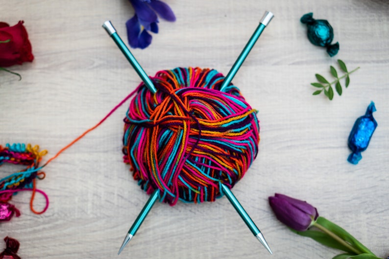 Pu&ograve; includere: Un gomitolo di lana colorata con due ferri da maglia turchesi, una rosa rossa, un fiore viola e due caramelle blu avvolte su una superficie di legno.