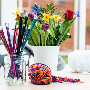 Pu&ograve; includere: Un barattolo di vetro pieno di ferri da maglia colorati, un gomitolo di lana multicolore, un gomitolo di lana bianco e un mazzo di fiori in un vaso bianco.