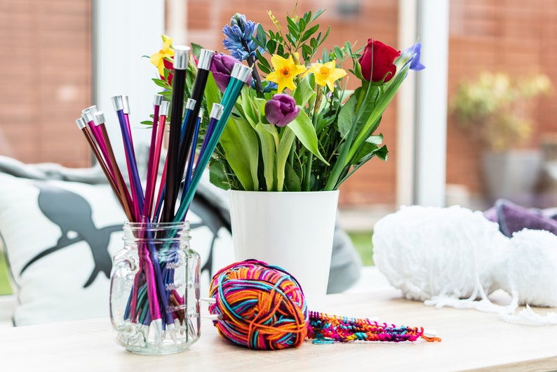 Pu&ograve; includere: Un barattolo di vetro pieno di ferri da maglia colorati, un gomitolo di lana multicolore, un gomitolo di lana bianco e un mazzo di fiori in un vaso bianco.