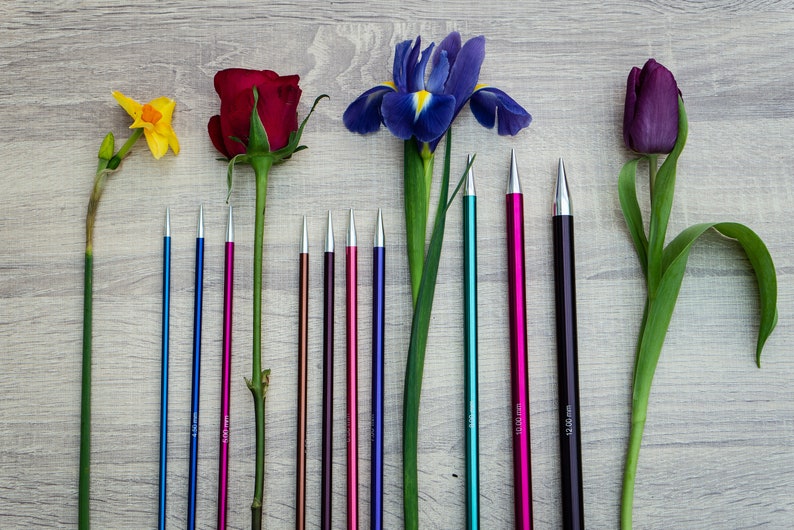 Pu&ograve; includere: Un set di 10 ferri da maglia di vari colori e dimensioni, che vanno da 3,5 mm a 10,0 mm, disposti accanto a fiori, tra cui un narciso giallo, una rosa rossa, un iris viola e un tulipano viola.