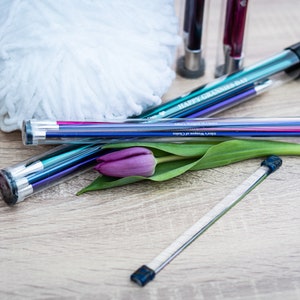 Pu&ograve; includere: Un set di ferri da maglia colorati in una custodia di plastica trasparente con un tappo nero. La custodia ha la scritta "Happy Birthday" stampata. I ferri sono accanto a un gomitolo di lana bianco, una rosa rossa e un tulipano viola.